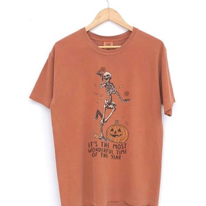 Könnte beinhalten: Zwei T-Shirts mit einem Skelett-Motiv und dem Text "It's the most wonderful time of the year". Das Shirt links ist orange und das Shirt rechts ist blau.