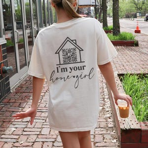 May include: Cream-colored t-shirt with a black house graphic and a QR code. The text "I'm your home girl" is printed below the graphic. The shirt is worn by a person walking on a brick walkway.