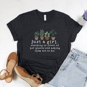 May include: A dark gray t-shirt with a white graphic of potted plants and the text "just a girl.. standing in front of her plants and asking them not to die."