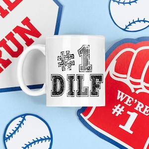 May include: White ceramic mug with black text that reads "#1 DILF". The mug is surrounded by baseball-themed decorations, including a red and white baseball, a red and white foam finger, and a red and white baseball sign that reads "HOME RUN".