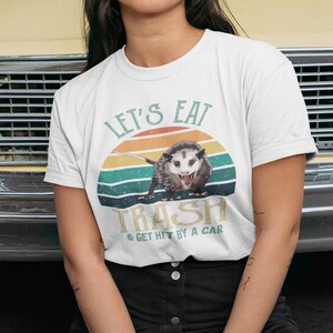 May include: A white t-shirt with a retro-style graphic of a possum with the text "Let's Eat Trash & Get Hit By A Car".