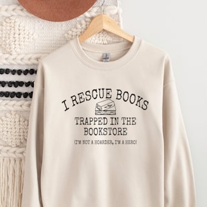 Może przedstawiać: Beżowa bluza z napisem "I rescue books trapped in the bookstore (I'm not a hoarder, I'm a hero)" i czarno-białą ilustracją ułożonych książek.