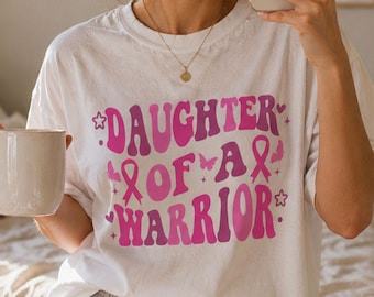 May include: A white short-sleeved t-shirt featuring the pink text DAUGHTER OF A WARRIOR in a wavy font. The design includes pink breast cancer awareness ribbons, small butterflies, and star icons. The shirt is paired with grey drawstring shorts.