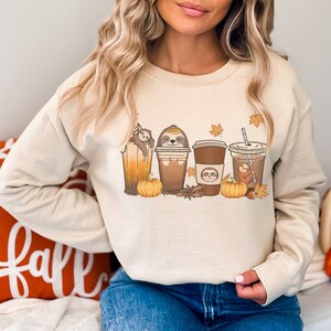 May include: A beige sweatshirt featuring a design of sloths in coffee cups and pumpkins. A pillow with the word "Fall" is visible in the background.