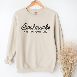 May include: A beige sweatshirt with the text "Bookmarks are for Quitters" in black script. The sweatshirt is hanging on a wooden hanger against a white background. A bundle of dried beige grass is to the right of the sweatshirt.