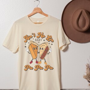 May include: A cream-colored t-shirt with a retro-style graphic of two pie slices with faces and the text "Ain't No Lie Baby Pie Pie Pie".