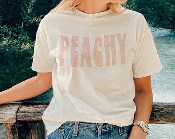 Op de afbeelding: Een wit T-shirt met het woord "PEACHY" in roze op de voorkant. Het shirt wordt gedragen door iemand die een spijkerbroek draagt.