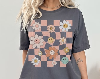 May include: A dark grey t-shirt featuring a central graphic of a wavy, checkered pattern in light pink and dark grey. The design is decorated with various flowers and smiley faces in shades of white, yellow, orange, and light green with black outlines.