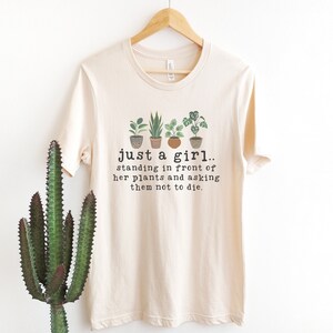 May include: A beige t-shirt with a graphic of potted plants and the text "just a girl.. standing in front of her plants and asking them not to die."