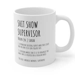 Könnte beinhalten: Weiße Keramik-Kaffeetasse mit einem schwarzen Design, auf dem steht: "Shit Show Supervisor Substantiv (N.) Sarah 1. Ein Problemlöser, der Probleme behebt, die niemand sonst lösen kann. 2. Eine Person, deren Aufgabe es ist, das Chaos zu organisieren, das regelmäßig entsteht Siehe auch: Wunderarbeiter, Superheld"