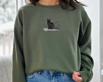 May include: A green sweatshirt with a black cat sitting on a book with flowers embroidered on it.