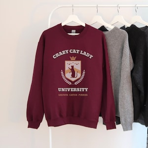May include: Burgundy sweatshirt with a graphic of a cat wearing a crown and a shield with the text "Crazy Cat Lady University" and the words "MEOWS - CATIUS - PURRIES" underneath.