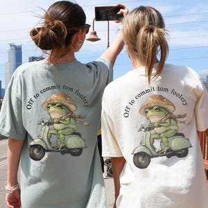 Peut inclure: Deux t-shirts amples, un de couleur baie et l'autre ivoire, avec un motif d'une grenouille sur un scooter et le texte "Off to commit tom foolery". La grenouille porte un chapeau et une écharpe. Les t-shirts sont portés par deux personnes sur un pont.