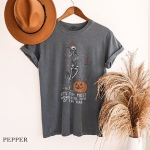 Könnte beinhalten: Ein dunkelgraues T-Shirt mit einem Skelett, das auf einem Kürbis tanzt. Der Text "It's the most wonderful time of the year" ist unter dem Bild gedruckt.