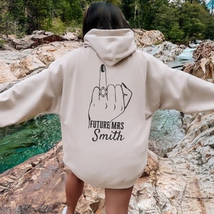May include: A beige hoodie with a black graphic of a hand giving the middle finger with a diamond ring on the ring finger. The text "FUTURE MRS Smith" is printed below the graphic.