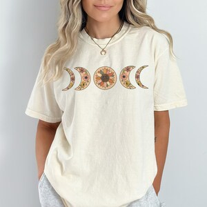以下が含まれることがあります： クリーム色のTシャツ。花の月の満ち欠けのデザインが特徴です。オレンジ、黄色、茶色の花で満たされたひまわりと三日月が描かれています。半袖でゆったりとしたフィット感です。