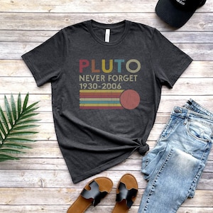 May include: A dark grey t-shirt with a vintage-style graphic that says "Pluto Never Forget 1930-2006" with a red circle representing Pluto. The shirt is laid out on a wooden surface with a pair of brown sandals, blue jeans, and a black baseball cap.