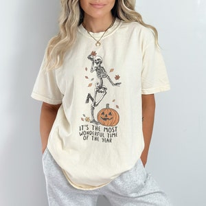 Könnte beinhalten: Ein cremefarbenes T-Shirt mit einem Skelett, das auf einem Kürbis tanzt. Der Text auf dem Shirt lautet "It's the most wonderful time of the year".
