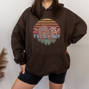 May include: A brown hoodie with a vintage-style graphic of a sloth sleeping on a branch in front of a sunset. The graphic is in shades of orange, yellow, red, and green.