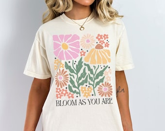 Puede incluir: Una camiseta blanca con un diseño floral y el texto "Bloom as you are".