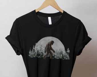 Może przedstawiać: Czarny T-shirt z grafiką przedstawiającą Bigfoota idącego przed pełnią księżyca i drzewami.