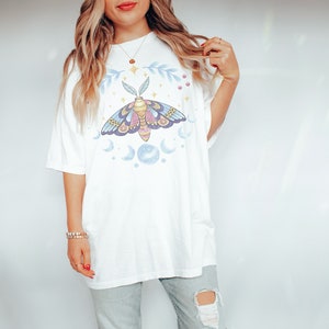 May include: White oversized t-shirt with a graphic of a moth with blue and purple wings, surrounded by moons and stars.