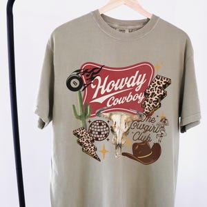 May include: Khaki-colored t-shirt with a graphic design featuring a steer skull, cowboy hat, and the words "Howdy Cowboy" in a red banner. Additional graphics include a cactus, lightning bolts, and an 8-ball.