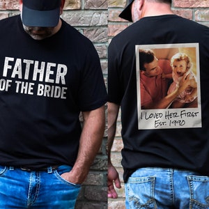 May include: Black t-shirt with the text "Father of the Bride" on the front and a photo of a father and daughter with the text "I Loved Her First Est. 1990" on the back.