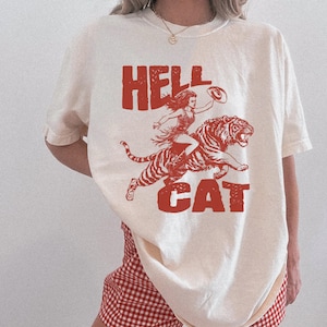 Könnte beinhalten: Ein weißes T-Shirt mit einem roten Aufdruck einer Frau, die auf einem Tiger reitet. Der Aufdruck lautet "Hell Cat".