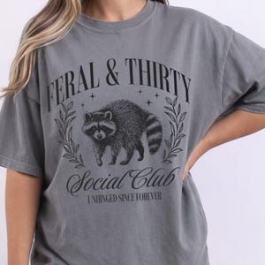 May include: A grey t-shirt with the text "FERAL & THIRTY Social Club" and a raccoon illustration. The design is in black and features a raccoon surrounded by leaves. The shirt is a relaxed fit.