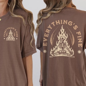 Może przedstawiać: Brązowy t-shirt z grafiką przedstawiającą szkielet siedzący w pozycji lotosu z płomieniami za nim. Tekst "EVERYTHING'S FINE" jest napisany w kółku wokół szkieletu.