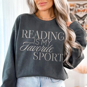 May include: A dark gray sweatshirt with the text "READING IS MY FAVORITE SPORT" in a light gray script. The sweatshirt is a crew neck style and is worn with light blue denim shorts. The word "Pepper" is in the bottom right corner.