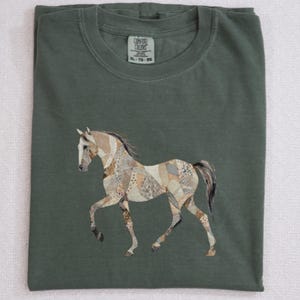 May include: Moss green t-shirt with a horse graphic. The horse is depicted with a patchwork design in shades of beige, brown, and gray. The shirt has a crew neck and short sleeves. The text "MOSS" is at the bottom right.
