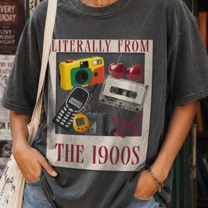 以下が含まれることがあります： LITERALLY FROM THE 1900Sという文字がプリントされたチャコールグレーの半袖Tシャツ。デザインには、黄色いカメラ、2つの赤いサクランボ、カセットテープ、黒い携帯電話、黄色いデジタルペット、蝶が描かれています。