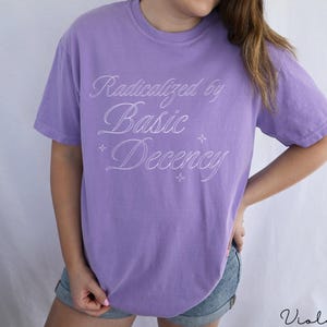 May include: A violet t-shirt featuring the white script text "Radicalized by Basic Decency". The shirt has a crew neck and short sleeves. The word "Violet" is written in script at the bottom right corner.