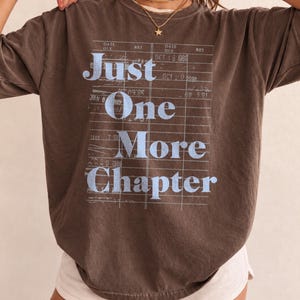 May include: A dark brown short-sleeve t-shirt featuring a light blue graphic print of a library due date card. The text reads Just One More Chapter, overlaid on a grid pattern with various dates including Oct 18 1989 and Apr 20 1988.