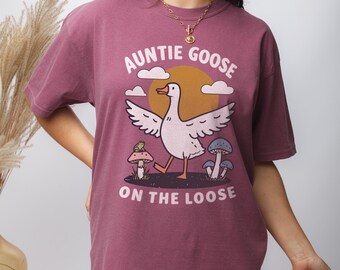 Może przedstawiać: Maronowy t-shirt z kreskówkową gęsią i napisem "Auntie Goose on the Loose".