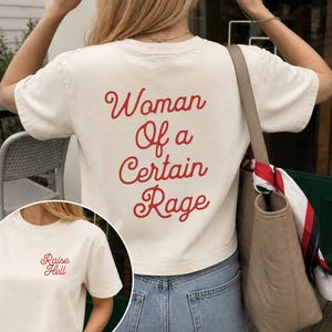 May include: An off-white short-sleeve t-shirt featuring red cursive text on the back that reads Woman Of a Certain Rage. The front of the shirt displays the red cursive text Raise Hell on the upper left chest area. The shirt is paired with blue jeans.