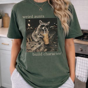 May include: Dark green t-shirt with a graphic of a raccoon wearing sunglasses and drinking iced coffee. The text "weird aunts build character" is printed above and below the raccoon image. The shirt has a relaxed fit.