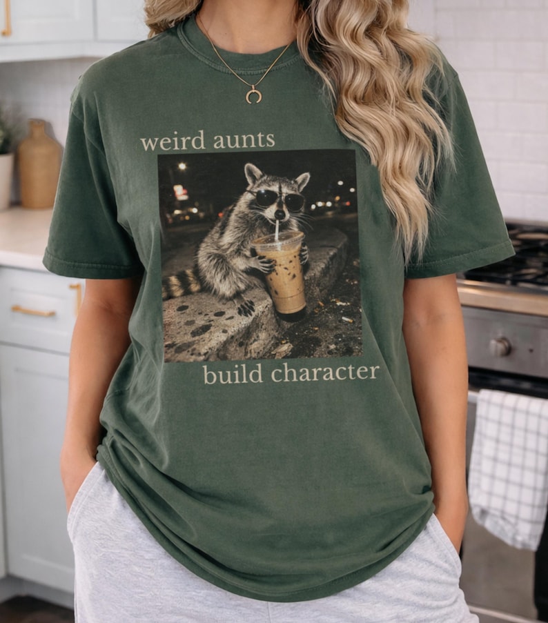 May include: Dark green t-shirt with a graphic of a raccoon wearing sunglasses and drinking iced coffee. The text "weird aunts build character" is printed above and below the raccoon image. The shirt has a relaxed fit.