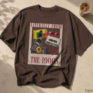 以下が含まれることがあります： LITERALLY FROM THE 1900Sというテキストがプリントされた長方形のグラフィックが中央にある、ダークブラウンの半袖Tシャツ。デザインには、黄色と赤のフィルムカメラ、黒い携帯電話、カセットテープ、サクランボ、黄色いデジタルペットが描かれています。
