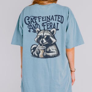 May include: A light blue t-shirt with a graphic of a raccoon wearing sunglasses and drinking from a cup. The text "CAFFEINATED AND FERAL" is printed on the shirt.