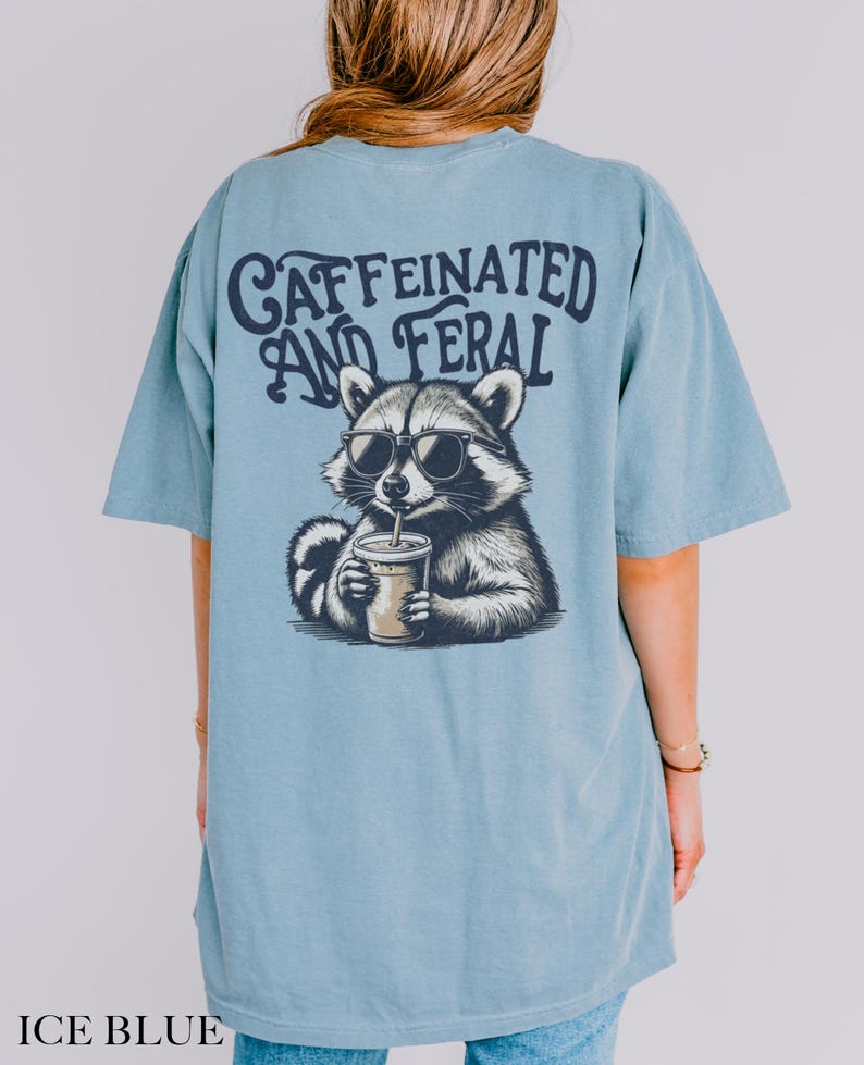May include: A light blue t-shirt with a graphic of a raccoon wearing sunglasses and drinking from a cup. The text "CAFFEINATED AND FERAL" is printed on the shirt.