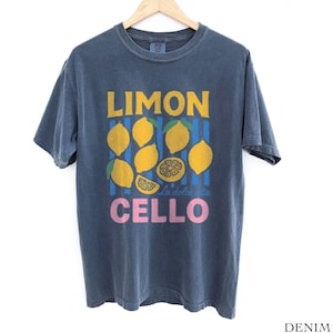 May include: Denim blue t-shirt with a lemon-themed graphic. The design features the word "LIMON" in yellow, lemons, blue stripes, and the word "CELLO" in pink. The shirt is hanging on a wooden hanger.