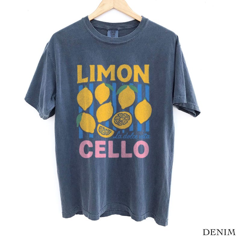 May include: Denim blue t-shirt with a lemon-themed graphic. The design features the word "LIMON" in yellow, lemons, blue stripes, and the word "CELLO" in pink. The shirt is hanging on a wooden hanger.