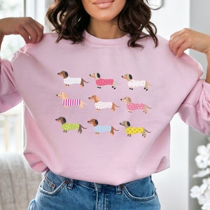 May include: A pink sweatshirt with a graphic print of multiple dachshunds wearing different patterned pajamas.