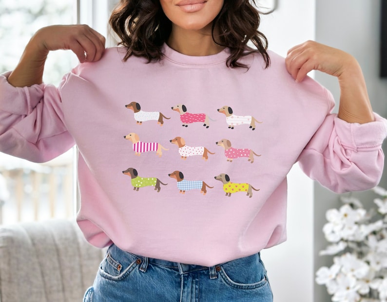 May include: A pink sweatshirt with a graphic print of multiple dachshunds wearing different patterned pajamas.