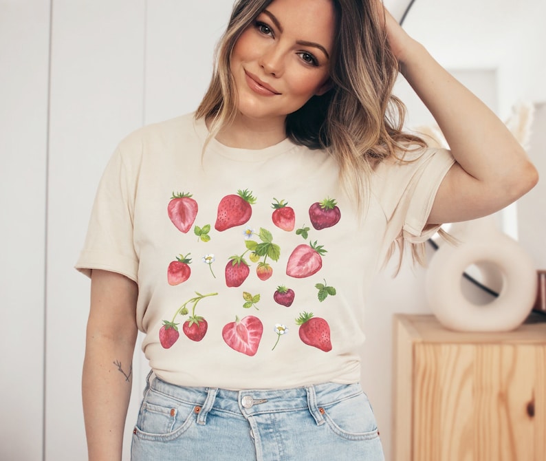 May include: A cream-coloured t-shirt with a pattern of red and pink strawberries, green leaves, and white flowers.