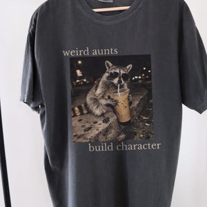 May include: Dark grey t-shirt with the text "weird aunts build character." The shirt features a graphic of a raccoon wearing sunglasses and drinking an iced coffee. The t-shirt is hanging on a wooden hanger.