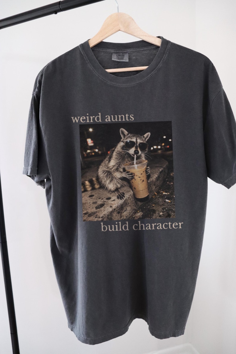 May include: Dark grey t-shirt with the text "weird aunts build character." The shirt features a graphic of a raccoon wearing sunglasses and drinking an iced coffee. The t-shirt is hanging on a wooden hanger.